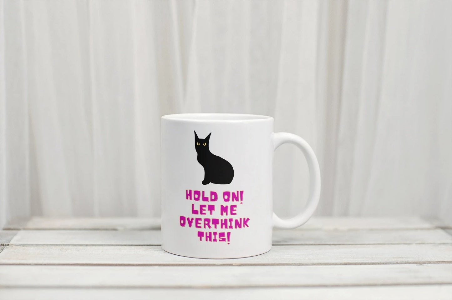 Niedliche weiße Tasse auf der eine ernst blickende schwarze Katze zu sehen ist. Darunter befindet sich ein pinker Schriftzug "Hold on! Let me overthink this!"